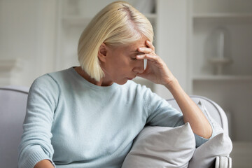 Unhappy depressed thoughtful mature woman touching head, sitting alone on couch at home close up, thinking about problems, sad stressed older female suffering from grief loneliness, mental troubles