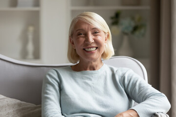 Fototapeta na wymiar Head shot portrait mature woman with healthy toothy smile, beautiful older female with grey hair looking at camera, sitting on cozy sofa at home, posing for photo, making video call, using webcam