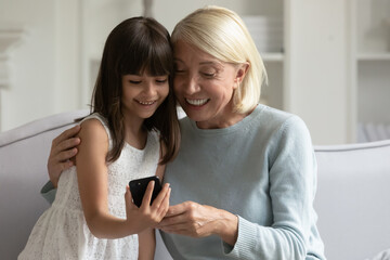 Happy grandmother and little granddaughter using phone together, having fun at home, sitting on cozy sofa, smiling positive senior granny hugging cute girl grandchild, playing game, browsing apps