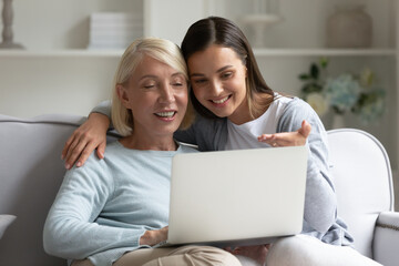 Happy mature mother and adult daughter using laptop, surfing internet or shopping online, older grandmother and granddaughter looking at computer screen, watching video or movie, having fun at home