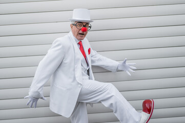 An elderly man in a white suit, huge boots and a clown nose walks funny. 