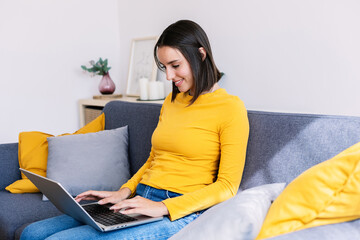 Joyful millennial female using laptop computer sitting on couch at apartment. Technology and...
