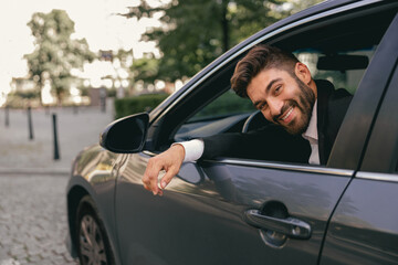 Smiling businessman in suit is riding behind steering wheel of car and looks camera