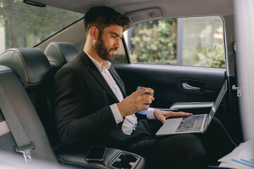 Professional male finansist working on laptop sitting car backseat and drinking coffee