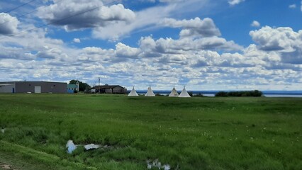 Beautiful shot of tipis set up on green plains, under a clear blue sky with scattered white clouds