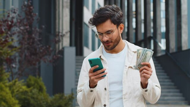 Happy rich young Indian man counting money dollar cash, use smartphone calculator app plans to order gifts and food delivery online, booking hotel room. Arabian Hindu guy in city street. Town outdoors
