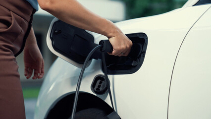 Progressive woman installs a charging station plug into her electric vehicle at home. EV automobiles provide an environmentally beneficial concept of clean and green energy. - obrazy, fototapety, plakaty