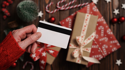 Hands of a woman in a red sweater with a credit card on a background of gift boxes, selective focus, close-up. Christmas and New Year shopping online, payment by credit card. Buying gifts online.