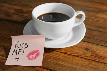 Sticky note with phrase Kiss Me and cup of coffee on wooden table