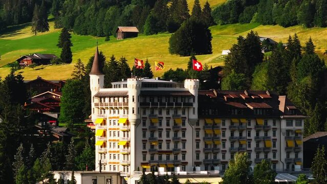 Zoomed view of luxurious hotel building with turrets and flags waving in wind. Picturesque colour mountain meadows in background. Gstaad, Switzerland