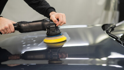 A mechanic polishes the surface of the hood of a gray car. 