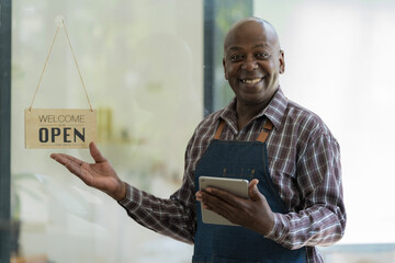 Portrait of an African barista, waiter cafe, or coffee shop owner against the entrance with an open signboard, a smiling guy in an apron standing and holding a digital tablet.