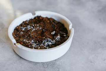 Dried coffee grounds and cigarette ash in a white ashtray. Copy space. White ashtray on gray ceramic table.