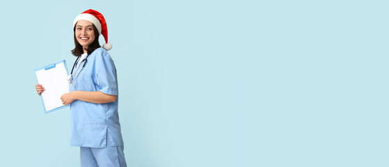 Female doctor in Santa hat holding clipboard on light blue background with space for text