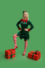Happy young woman in elf costume with Christmas gift boxes on green background