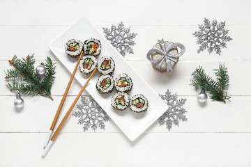 Plate of tasty sushi rolls with fir branches, gift box and Christmas decorations on white wooden background