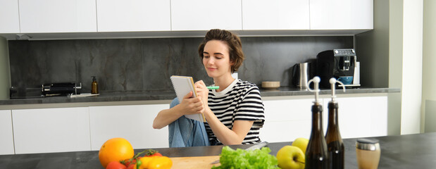 Portrait of beautiful modern woman, girl cooking with vegetables, holding notebook, writing down...