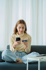 Obraz premium Cheerful Asian woman using smartphone, shopping online, holding credit card, paying, sitting on sofa at home Internet banking and e-commerce applications vertical picture