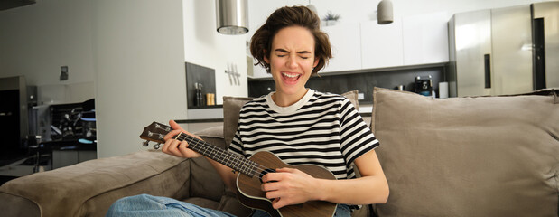 Beautiful girl playing ukulele on sofa, singing her favourite song and picking chords on small...