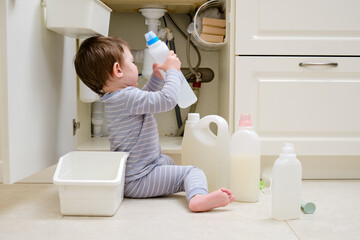 A child is playing with chemical cleaning products under the sink in the kitchen. Baby holds bottles with detergent. Kid aged about two years (one year nine months) - obrazy, fototapety, plakaty