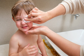 A woman mother cleans her child eyes with a cotton pad in the home bathroom. Kid aged two years (two-year-old boy) Mom washes toddler baby eyes