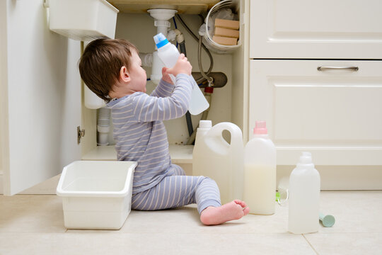 A child is playing with chemical cleaning products under the sink in the kitchen. Baby holds bottles with detergent. Kid aged about two years (one year nine months)