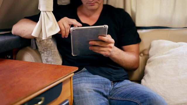 A young guy is relaxing on the sofa in a motor home watching video on his tablet and smiling