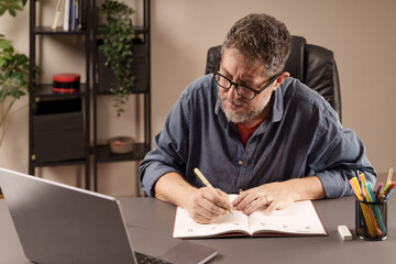 Man focused on writing in his planner, organizing tasks while working on his laptop in a home...