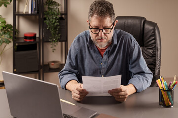 Concentrated mature man reading a paper document at his home office desk, engaged in work-related...
