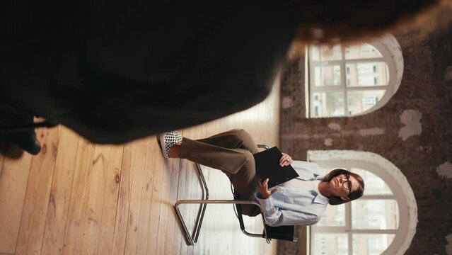 Vertical video: a girl psychologist with a bob hairstyle and glasses in a blue shirt communicates with a participant in group therapy and interviews him