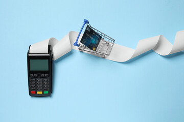 Payment terminal with thermal paper for receipt, small shopping cart and credit card on light blue background, flat lay