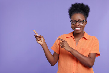 Happy young woman in eyeglasses pointing at something on purple background. Space for text