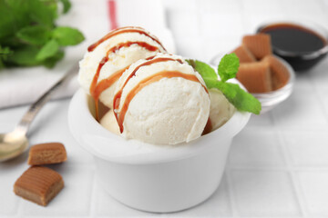 Scoops of tasty ice cream with mint leaves, candies and caramel sauce on white tiled table, closeup