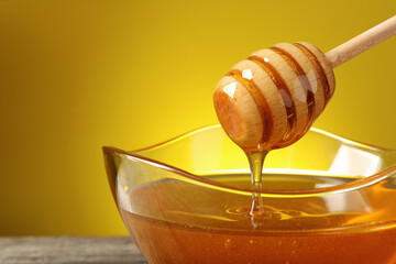 Delicious honey flowing down from dipper into bowl on table against yellow background, closeup....