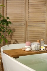 Wooden tray with spa products, stones and burning candle on bath tub in bathroom. Space for text