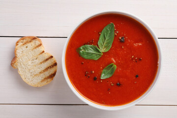 Delicious tomato cream soup in bowl and piece of grilled bread on white wooden table, flat lay