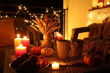 Rattan furniture, cups, fairy lights, burning candles and other autumn decor on outdoor terrace at night