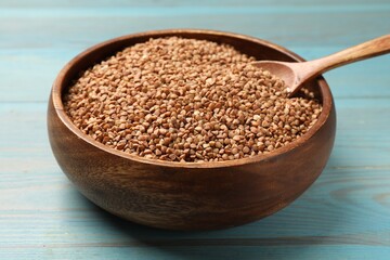 Bowl and spoon with dry buckwheat on light blue wooden table, closeup