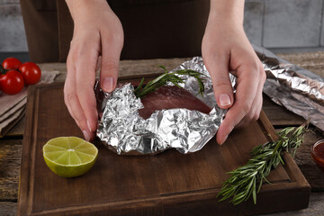 Woman wrapping meat in aluminum foil at wooden table, closeup