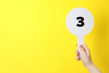 Woman holding auction paddle with number 3 on yellow background, closeup. Space for text