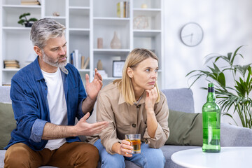Upset couple sitting in living room on sofa, alcoholic woman drinking a lot of strong alcohol alone, man trying to stop her, discuss everything, relationship problems, alcohol addiction. © Liubomir