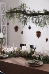 Christmas celebration. Cones hanging from fir tree branch over table with burning candles and tableware