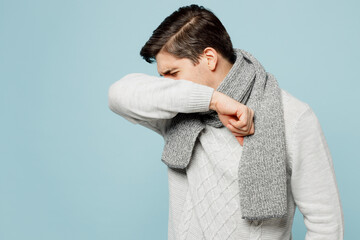 Young ill sick man wear gray sweater scarf cough cover mouth with hand sleeve isolated on plain blue background studio portrait. Healthy lifestyle disease virus treatment cold season recovery concept.