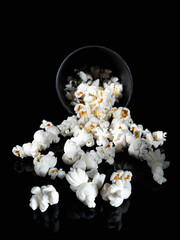 Frontal view of popcorn falling out of a black paper cup on a black background with reflection. Selective focus. Vertical shot. Isolate.