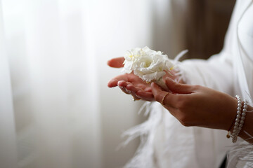 The bride is standing by the window, holding a wedding boutonniere in her hands. Preparation for the wedding ceremony. - obrazy, fototapety, plakaty