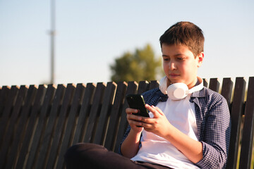 a cute boy teenager in headphones , watches mobile phone and sits on bench outdoor in the park on blue sky background.