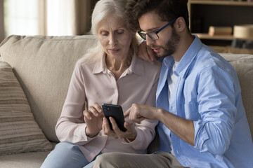 Middle aged woman spend time with grownup son at home, sit on sofa in living room use smartphone...