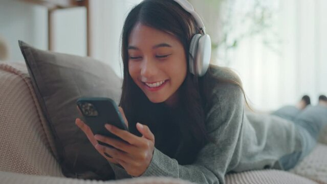 Happy young asian woman wear wireless headphones having fun listen music in smartphone and sit on sofa in living room, Female enjoys listening chill music audio sound feeling no stress at home.