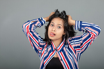 Portrait of a young Indian woman wearing black top and red, blue, white striped tunic, isolated on grey background. Young female model grabbing her hair showing happy and surprised expression