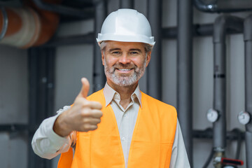 senior gray haired Smiling satisfied worker engineer in working clothes, with helmet and hardhat standing in factory and showing okay sign. thumb up in boiler room